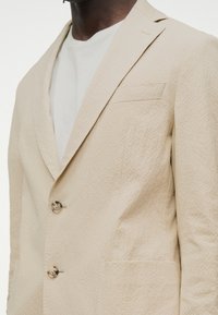 Άνδρας που φοράει μπεζ σακάκι με υφή, με γιακά notch και δύο κουμπιά, πάνω από λευκό πουκάμισο, κομμένο στον λαιμό και στη μέση του κορμού.
