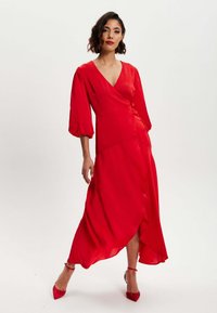 Red, flowy dress with a deep V-neck, three-quarter balloon sleeves, and an asymmetrical hem. Smooth fabric with a loose fit.