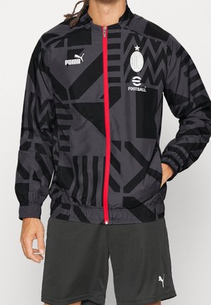 Czarna i szara kurtka na zamek błyskawiczny z geometrycznym wzorem, czerwonym zamkiem, logo futbolowe AC Milan oraz marką Puma, noszona z czarnymi szortami.