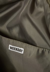 Olive green fabric with a smooth texture, featuring a small white label with the text "WEEKDAY" stitched onto a pocket edge.