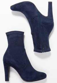 Par de botines de ante azul marino con cremallera lateral y tacones altos en fondo blanco.