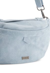 Lichtblauwe suède tas met een ronde vorm, voorzien van een bovenflap en een gemerkte metalen tag. Verstelbare schouderband met metalen hardware accenten.