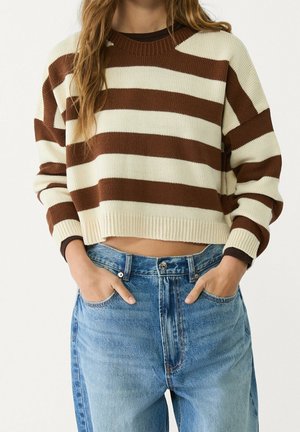 Vrouw draagt een bruin-crème gestreepte cropped trui met lange mouwen en hoog getailleerde blauwe spijkerbroek, handen in de voorzakken.