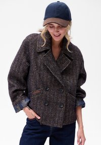 Kobieta ubrana w podwójnie zapinaną kurtkę w brązowo-niebieski jodełkowy wzór, ciemnoniebieskie dżinsy i granatową z brązowym czapkę baseballową, uśmiechnięta i patrząca w dół.