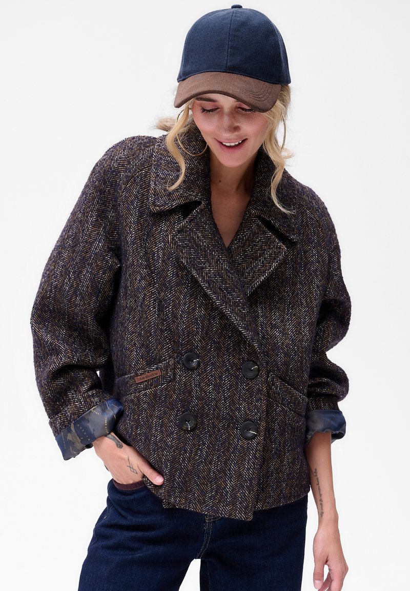 Kobieta ubrana w podwójnie zapinaną kurtkę w brązowo-niebieski jodełkowy wzór, ciemnoniebieskie dżinsy i granatową z brązowym czapkę baseballową, uśmiechnięta i patrząca w dół.