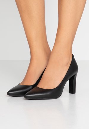 Tacones altos de cuero negro con punta en punta, usados sobre pies desnudos, de pie sobre una superficie blanca y lisa con un fondo neutro.