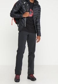 Černá izolovaná bunda se zipem vpředu, v kombinaci s tmavými kalhotami s texturovanými panely a turistickými botami s červenými akcenty. Batoh je součástí.