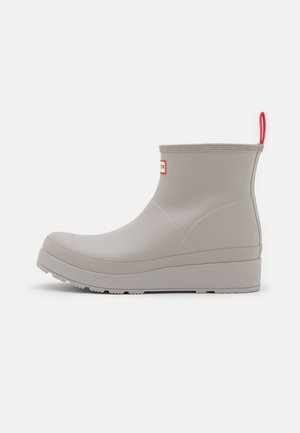 Matte beige ankle rain boot with thick sole and red pull tab on heel, side logo patch, and textured rim on a plain background.