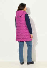 Lång ärmlös pufferväst i klar rosa med huva, med diamantstygn och en nederkant som faller under höfterna, i kombination med blå jeans.