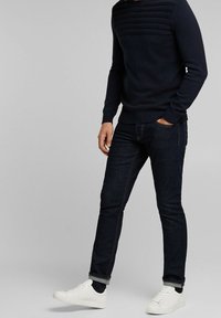 Marineblauwe gebreide trui met horizontale ribbels op de borst, gecombineerd met donkere spijkerbroek en witte sneakers, met een aansluitend ontwerp.