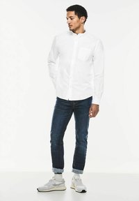 Biała koszula z długim rękawem i stójką, z kieszonką z przodu, połączona z niebieskimi dżinsami i jasnoszarymi tenisówkami z białymi akcentami.