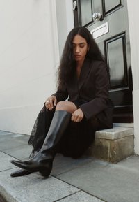 Bottes hautes jusqu'au genou en cuir noir avec des bouts pointus, portées avec une jupe en dentelle noire et un blazer sombre, posées contre une marche en pierre et une porte noire.