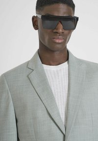 Blazer grigio chiaro con finitura testurizzata, maglione bianco a coste sotto, e occhiali da sole neri oversize e angolari.