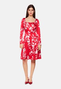Rotes Blumenkleid mit langen Ärmeln, quadratischem Ausschnitt und schwingendem Rock. Es hat weiße Blumenmuster auf dem leuchtend roten Stoff.