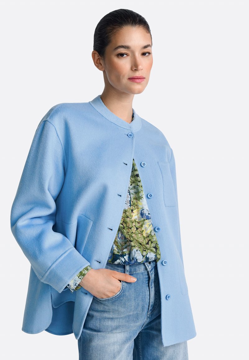 Chaqueta azul claro de mezcla de lana con corte recto, cuello redondo, botones frontales y bolsillos laterales. Combinada con una blusa de estampado floral.