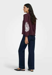 Mørkerød bluse med hvide broderede blomster mønstre på ærmerne, parret med mørkeblå brede jeans og sorte slip-on sko.