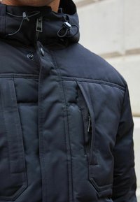 Čierna vypchatá bunda s vysokým golierom, nastaviteľnými šnúrkami a zipsovým vreckom. Vyrobená z hladkej, odolnej tkaniny.