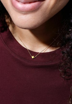 Zarte Goldkette mit einem kleinen runden Anhänger, getragen über einem maronenfarbenen Rundhalsausschnitt-Shirt. Glatte Textur und subtiler Glanz.