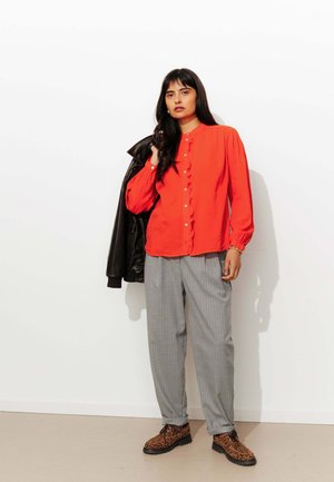 Chemise rouge à boutons avec un plastron froncé, pantalon gris à rayures, et chaussures brunes à imprimé léopard ; le modèle tient une veste en cuir noire.