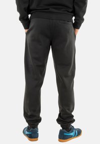 Pantalones de chándal negros, hechos de material suave, con una cinturilla elástica y puños, un bolsillo trasero y un corte ajustado. Combinados con zapatillas azules.