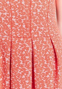 Una tela con un patrón floral rojo presenta manchas texturizadas y en relieve sobre un fondo rosa claro. El diseño incluye costuras estructuradas y pliegues.