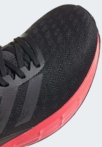 Černá sportovní bota s texturovaným pleteným svrškem, černými detaily a živým růžovým mezipodešví. Obsahuje černé šněrování a branding se třemi pruhy.