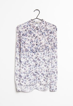 Blouse blanche à manches longues avec un motif floral bleu et rose, boutonnée à l'avant, avec un col classique, suspendue à un cintre noir sur un fond blanc.