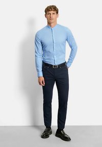 Lichtblauw overhemd met lange mouwen en een kraag zonder punten, met knopen aan de voorkant, gecombineerd met donkerblauwe broek en zwarte schoenen, staand tegen een neutrale achtergrond.