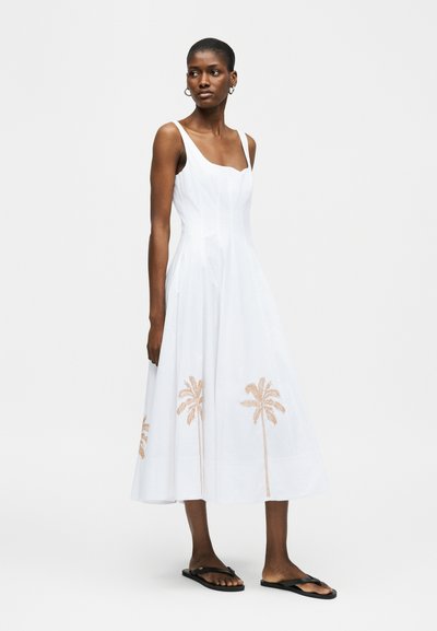Γυναίκα με λευκό midi φόρεμα χωρίς μανίκια, με κέντημα από φοίνικες κοντά στο στρίφωμα, συνδυασμένο με μαύρες επίπεδες σανδάλες.