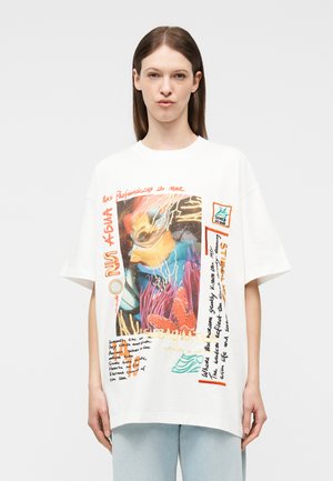 Kvinde iført en hvid, oversized t-shirt med farverigt, undervandstema grafik og tekst, kombineret med lyseblå jeans, neutral baggrund.