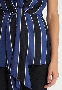 Blå och svartrandig topp med knyt framtill, tillverkad av ett mjukt och lätt material, med tunna vita ränder som framträdande detaljer.