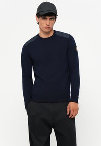 Suéter azul marino con mangas largas, detalles texturizados en los hombros, puños y cintura acanalados. Combinado con una gorra negra.