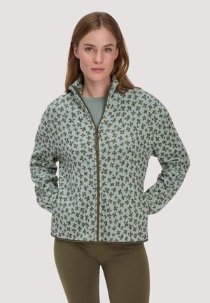 Vrouw draagt een lichtblauwe fleecejas met bruine klaverpatronen, dichtgeritst, met handen in de zakken, gecombineerd met olijfgroene broek.