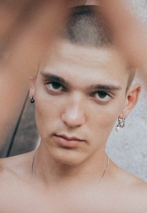 Silberne Schmuckstücke schmücken eine Person mit kurzen Haaren, die Ohrstecker in Form von Ringen und eine Halskette trägt, vor einem sanft strukturierten Hintergrund.