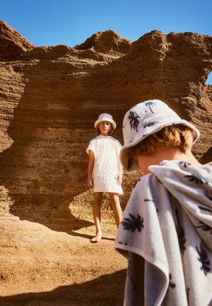 Δύο παιδιά σε ένα τοπίο ερήμου. Το ένα φοράει ένα λευκό κεντητό φόρεμα και ένα λευκό καπέλο του πλέι. Το άλλο έχει μια μοτίβο πετσέτα και ένα καπέλο του πλέι.