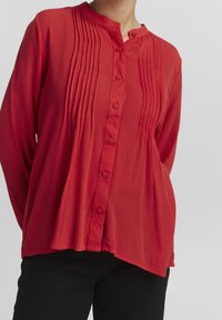 Camisa roja de botones con cuello alzado, con pliegues verticales en el frente, tejido texturizado y un corte relajado, combinada con pantalones negros.