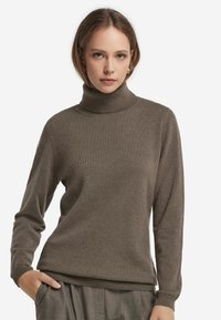 Ung kvinna som bär en brun polotröja och grå byxor, poserar med en hand i fickan mot en vit bakgrund.