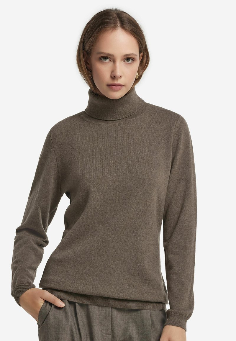 Ung kvinna som bär en brun polotröja och grå byxor, poserar med en hand i fickan mot en vit bakgrund.