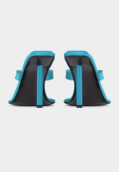 Ζευγάρι τιρκουάζ σανδάλια με ψηλά τακούνια, τετράγωνες μύτες, ένα φαρδύ λουράκι και κυλινδρικά τακούνια στιλέτο, εμφανιζόμενα από πίσω.