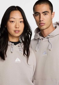 Des sweat-shirts beige clair avec des logos brodés, des cordons de serrage réglables et une texture douce. Portés par deux personnes, se tenant proches l'une de l'autre.