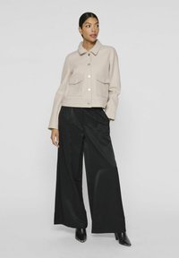Chaqueta beige corta con dos bolsillos frontales y botones plateados, combinada con pantalones anchos negros y botas de tacón negras. Textura suave.
