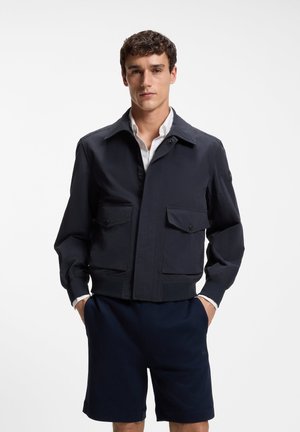 Giovane uomo che indossa una giacca blu scuro con grandi tasche frontali sopra una camicia bianca e pantaloncini blu, in piedi con le mani nelle tasche.