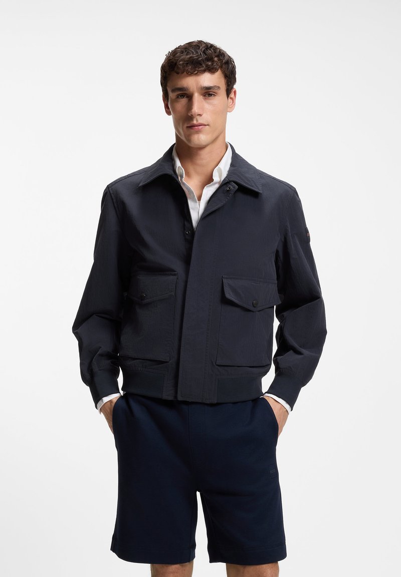 Jeune homme portant une veste bleu marine foncé avec de grandes poches devant, par-dessus une chemise blanche et un short bleu marine, debout les mains dans les poches.