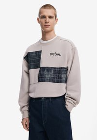 Light beige sweatshirt featuring a patchwork design with navy and black check patterns, embroidered logo, and ribbed cuffs and neckline.