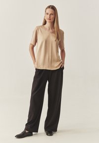 Beige Seiden-Shortsleeve-Top mit V-Ausschnitt, kombiniert mit locker sitzenden schwarzen Hosen und schwarzen Schuhen mit strukturierter Oberfläche.