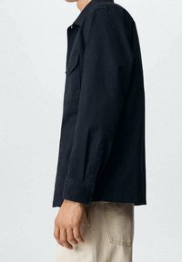 Ciemnoniebieska koszula z długim rękawem z bawełny, z kołnierzykiem, kieszenią z przodu i zapinanymi mankietami. Noszona z jasnymi spodniami.