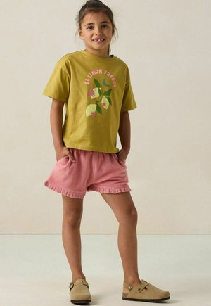 Grünes Baumwoll-T-Shirt mit floraler Grafik, kombiniert mit rosa Frottier-Shorts mit einem Rüschensaum. Beige Clogs vervollständigen das Outfit.