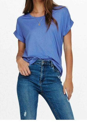 Mujer con camisa azul de manga corta metida por dentro con puños remangados y vaqueros azules de talle alto con ligero desgaste en los muslos.