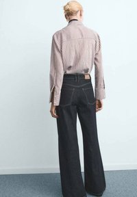 Camisa de rayas de manga larga en rojo y blanco, combinada con jeans de mezclilla oscuros y de pierna ancha. Incluye aberturas en las mangas y un detalle de etiqueta en la parte posterior.