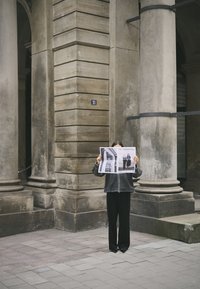 Eine Person hält ein Magazin mit dem Titel "DIE ESSENTIALS", trägt eine dunkelgraue Jacke und schwarze weite Hosen und steht in der Nähe eines mit Säulen versehenen Gebäudes.